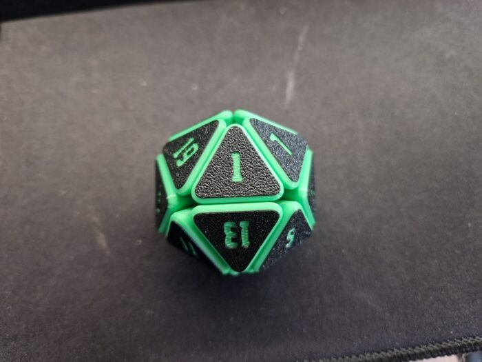 Grüner Squishy D20 Fidget Würfel auf Würfelmatte Grüner Squishy D20 Fidget Würfel mit schwarzen Flächen und grünen Zahlen, liegend auf einer dunklen Würfelmatte – ideal für D&D und Tabletop-Runden.
