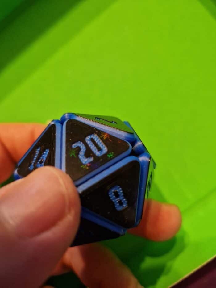 Blauer Squishy D20 Fidget Würfel mit Glitzereffekt in Hand „Nahaufnahme eines blauen Squishy D20 Fidget Würfels mit schimmerndem Glitzereffekt, gehalten in einer Hand auf grünem Hintergrund