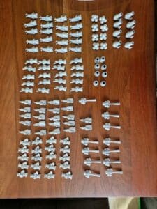 Mehrere Reihen von 3D-gedruckten Resin-Miniaturen und Zubehörteilen auf einem Holztisch. Ungrundierte Figuren und Spielmarker für Tabletop und Rollenspiele.